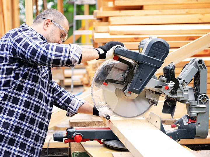 Een bouwvakker in een geruit overhemd en met een veiligheidsbril op, die met een verstekzaag een houten plank zaagt